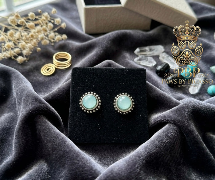 Pair of earrings on a black stand with silver and turquoise stones, surrounded by decorative items on a dark fabric background.