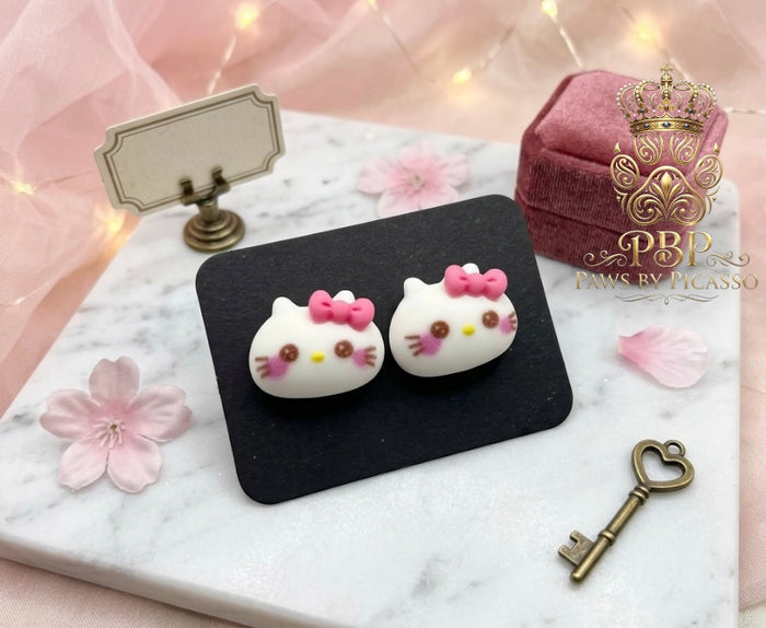 Cat-shaped earrings with pink bows on a black stand with decorative elements.