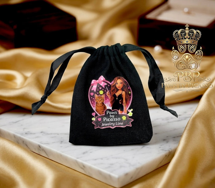 Black drawstring bag with 'Paws Picasso' jewelry line design on a marble surface with gold fabric background.