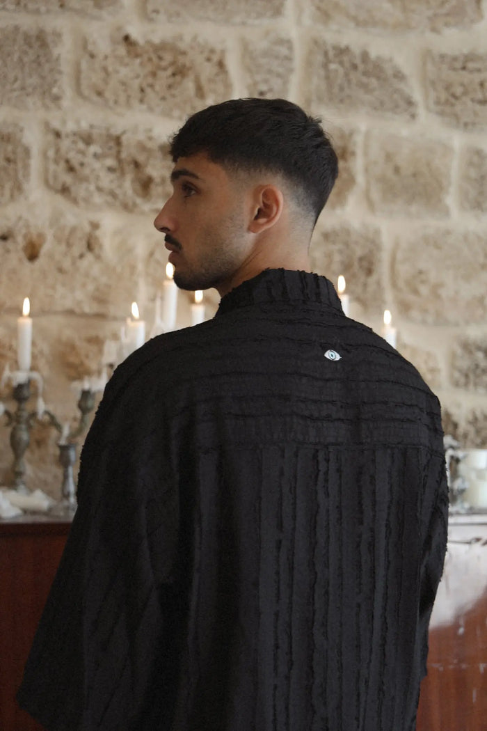 Man wearing a RIPS Short Sleeve Button-Up Shirt in black, facing away from the camera, looking left. A stone wall and lit candles are in the background