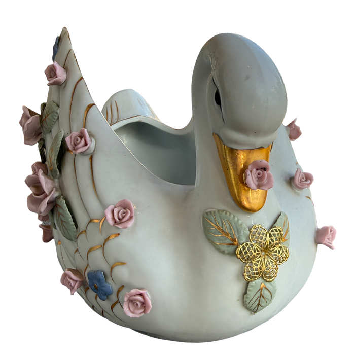 A decorative ceramic swan-shaped planter with a glossy off-white glaze, featuring a gold-painted beak, gold and green leaf details, and pink roses with blue accents on its body and tail.