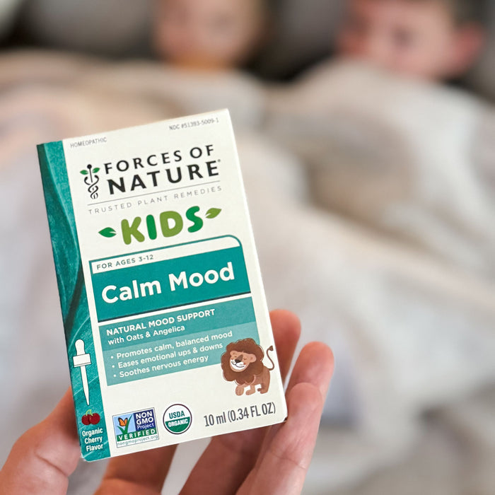 Front view of Calm Mood for Kids 10 ml box held in hand showing ‘Calm Mood’ label, natural mood support text, and USDA Organic and Non-GMO badges with a blurred bedroom background