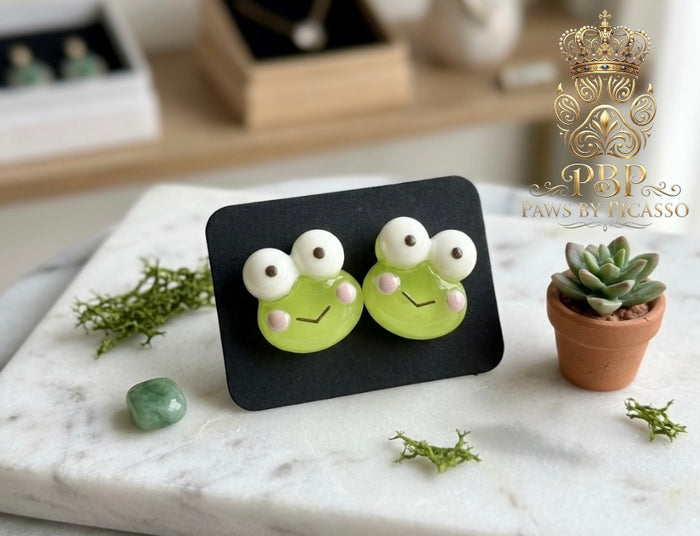 Frog-shaped earrings on a black stand with decorative elements on a marble surface.