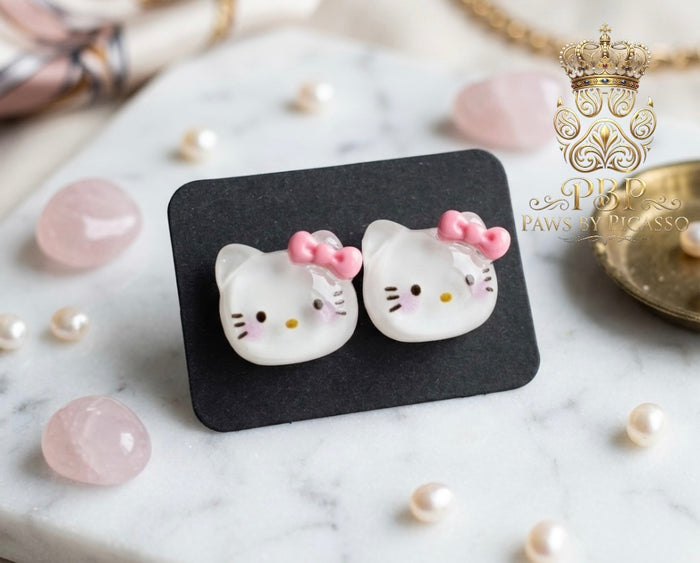 Cat-shaped earrings with pink bows on a black card, surrounded by pearls and gemstones.