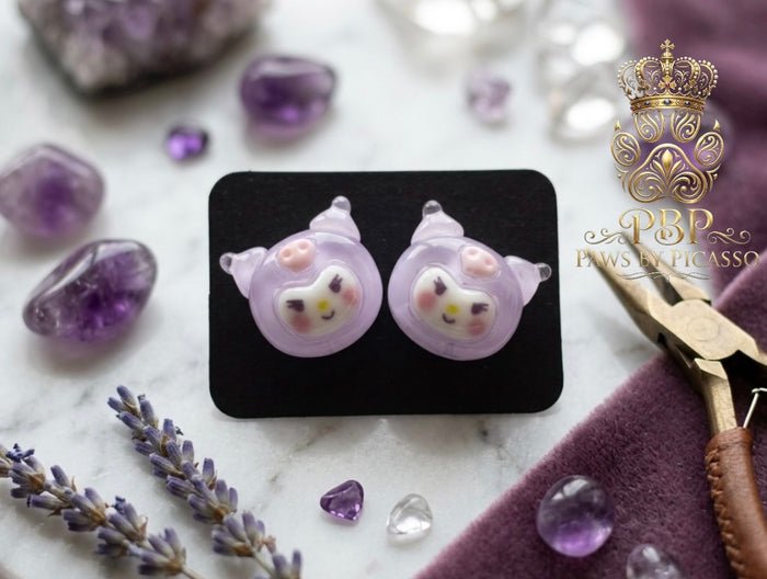 Pair of purple cat-shaped earrings on a black card with amethysts and lavender flowers in the background.