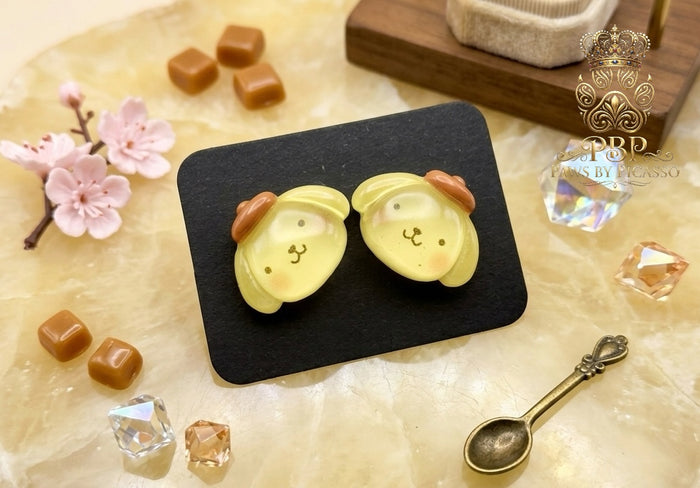 Two cute yellow dog-shaped earrings on a black square with decorative elements on a light brown surface.