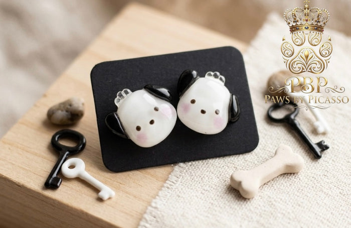 Pair of dog-shaped earrings on a black card with keys and a bone on a wooden surface, featuring the brand 'Paws by Picasso'.