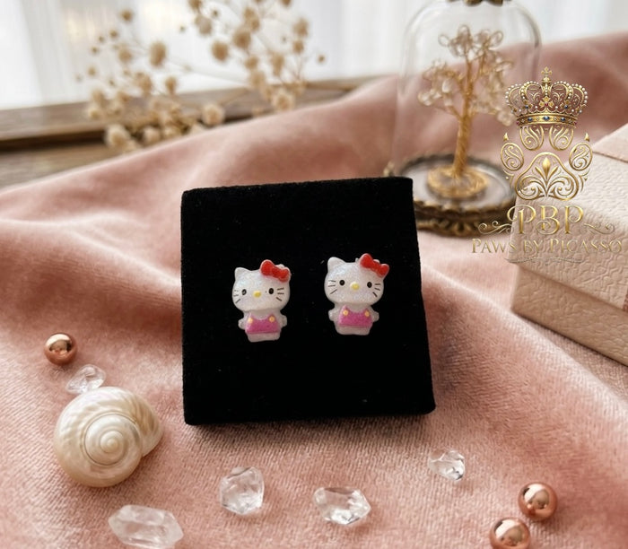 Cartoon cat earrings on a black stand with decorative elements on a pink surface