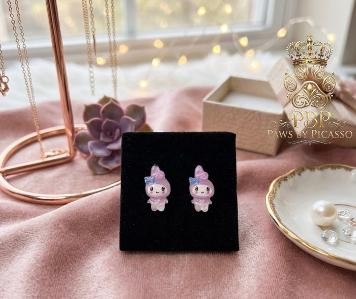 Pair of small animal-themed earrings on a black display card with a pink background and jewelry-related items.