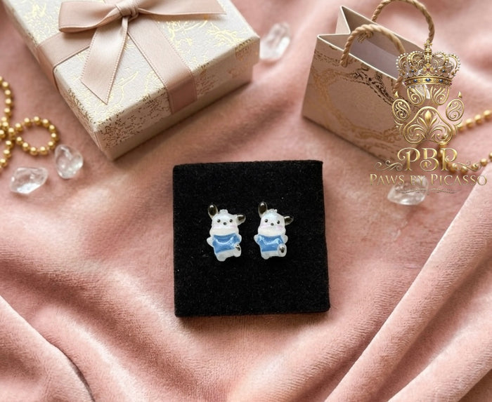 Pair of dog-shaped earrings on a black stand with a gift box and decorative items in the background.