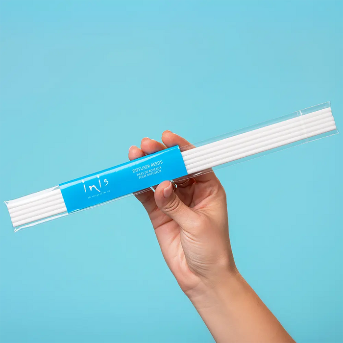 A hand holding a clear package of white reeds with a bright blue label that reads 'Inis' against a solid light blue background.