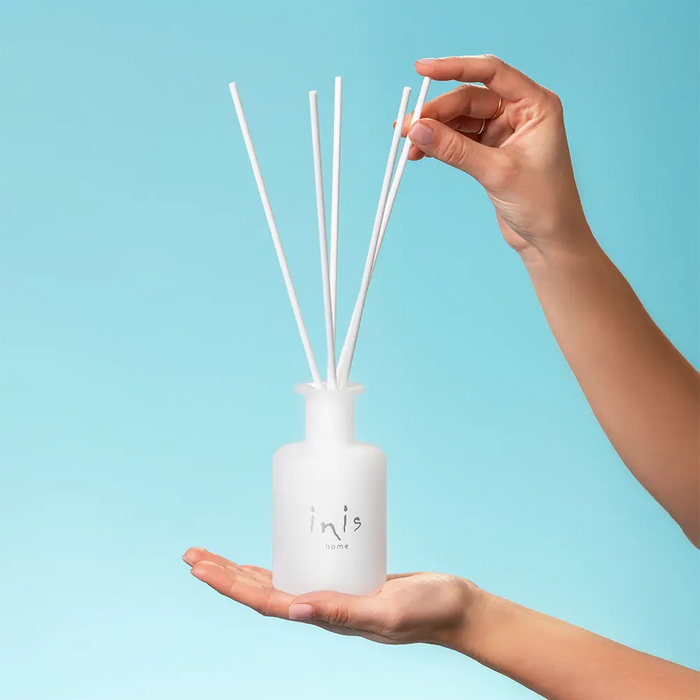 A hand holds a frosted glass reed diffuser bottle with white reeds, while another hand adjusts one of the reeds, against a solid light blue background. The bottle is branded with 'inis'.