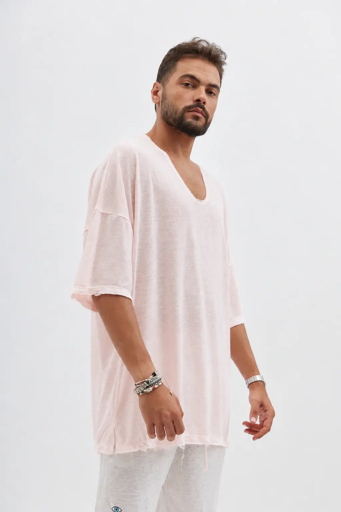 Man wearing a Dor Oversized Shirt in baby pink with a deep V-neck and distressed edges, paired with light grey trousers