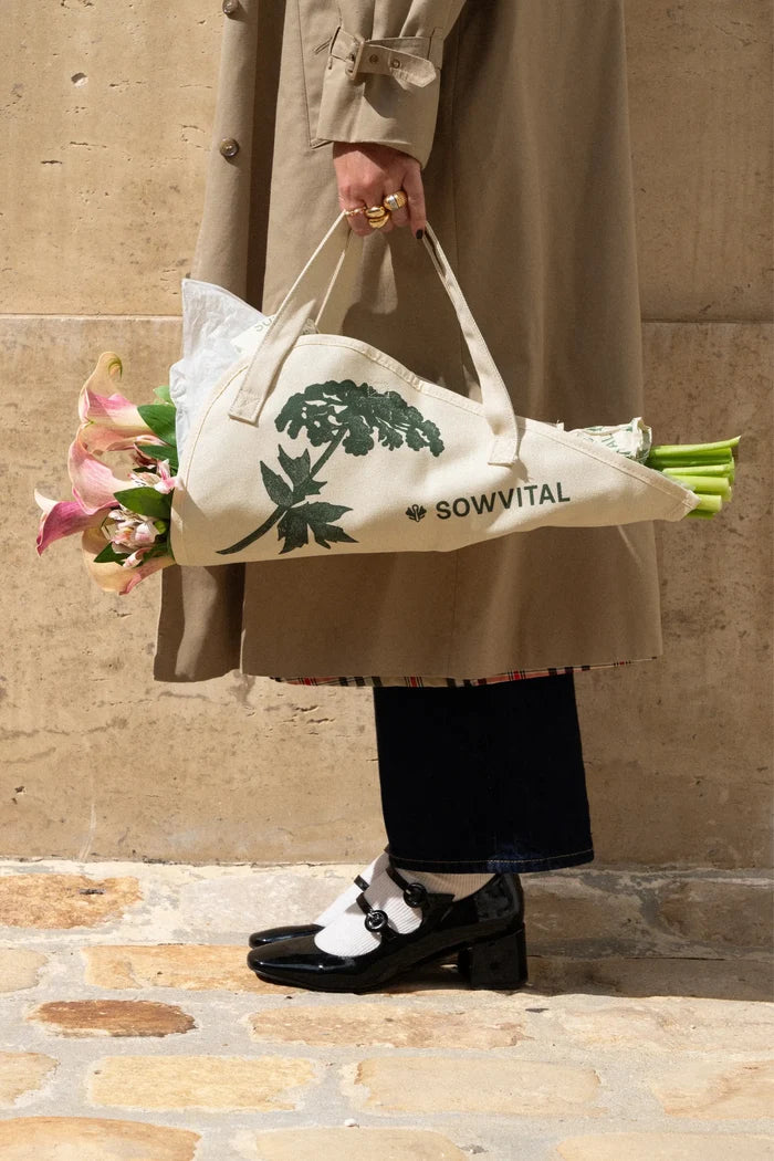 Flower Tote Monet Quote Classic. Person holding a white tote bag with green text and floral arrangement against a stone wall.