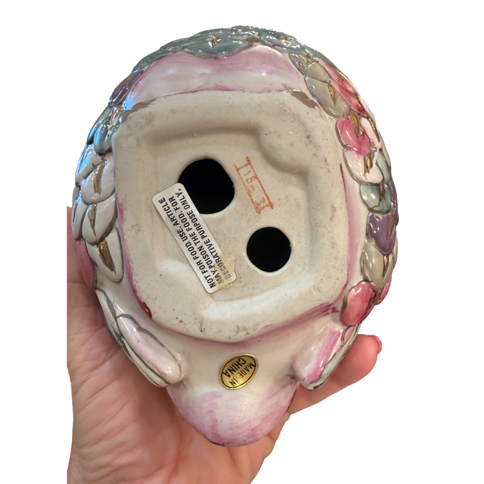 The underside of a vintage ceramic clown figurine held in a hand, showing the glazed pink and white face, decorative floral side details, and a white manufacturer label.