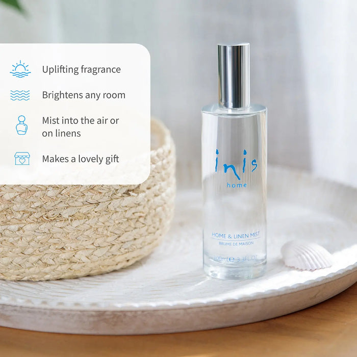 A clear glass bottle of inis home Home & Linen Mist with a silver cap, displayed on a white tray next to a woven straw hat and a small seashell.