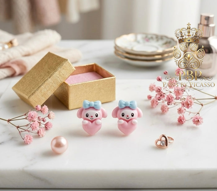 Pink animal-shaped earrings with a gold box and flowers on a marble surface, featuring the brand 'Paws By Picasso