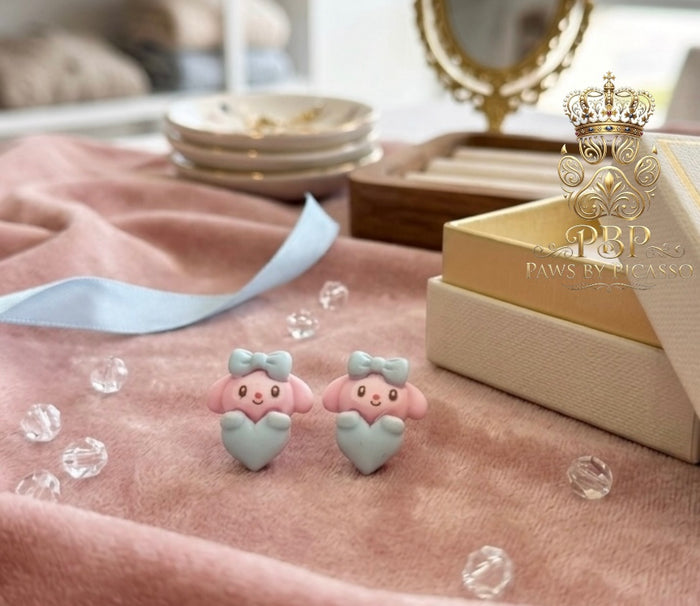 Pink animal-shaped earrings on a pink fabric background with a brand logo in the corner.