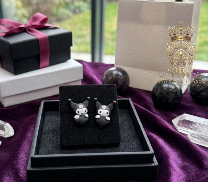 Black cat-shaped earrings on a black stand with a purple ribbon and gift box in the background.
