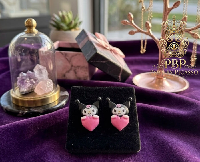 Pair of earrings with pink hearts in a black display case on a purple surface, with 'Paws by Picasso' branding.
