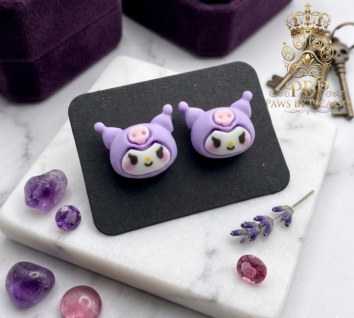 Purple cartoon-themed earrings on a black stand with decorative stones on a marble surface.