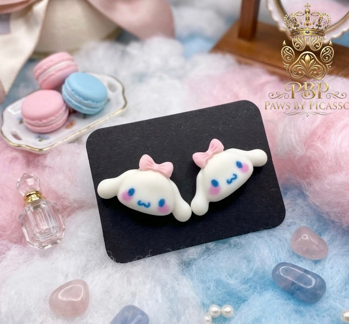 Pair of cartoonish cat-shaped earrings with pink bows on a black card, surrounded by decorative items.