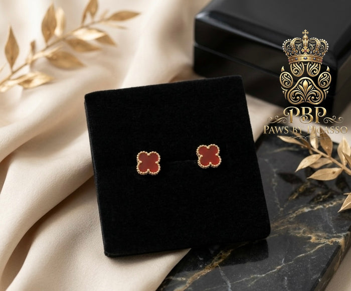 Clover-shaped earrings on a black velvet stand with a decorative box and leaves in the background, featuring the brand 'Paws by Picasso'.