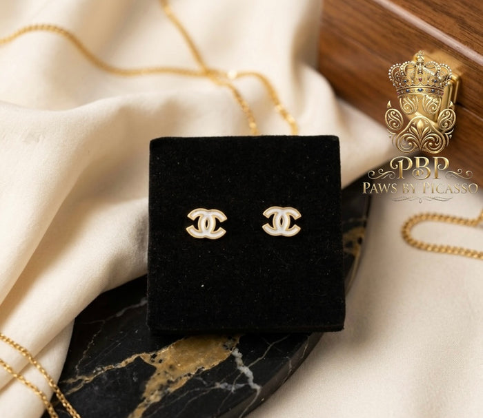 Gold Chanel earrings in a black display on a marble surface with a brand logo in the corner.