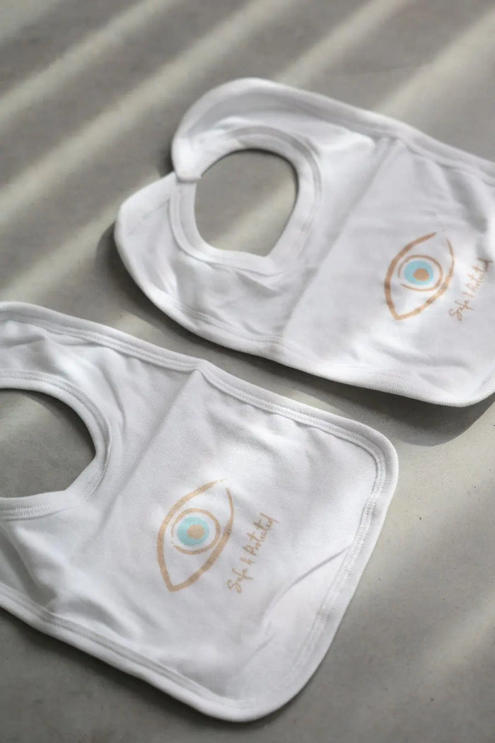Two white Newborn Cotton Bibs with an eye graphic and Safe & Protected text