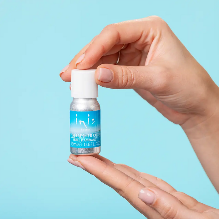 A hand holds a small bottle of inis refresher oil against a solid light blue background