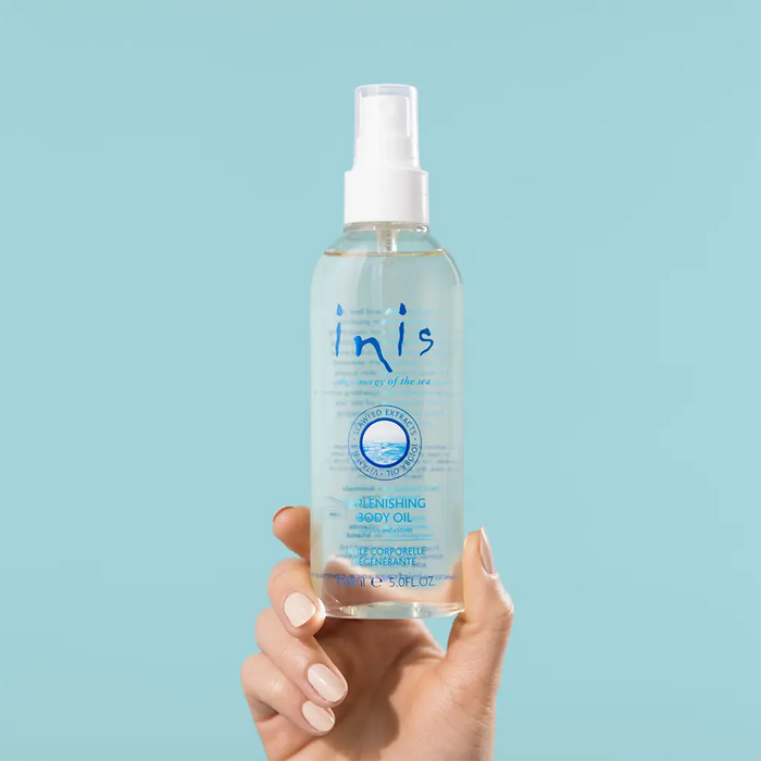 A hand with manicured nails holds a bottle of Inis Body Oil Spray against a solid light blue background.