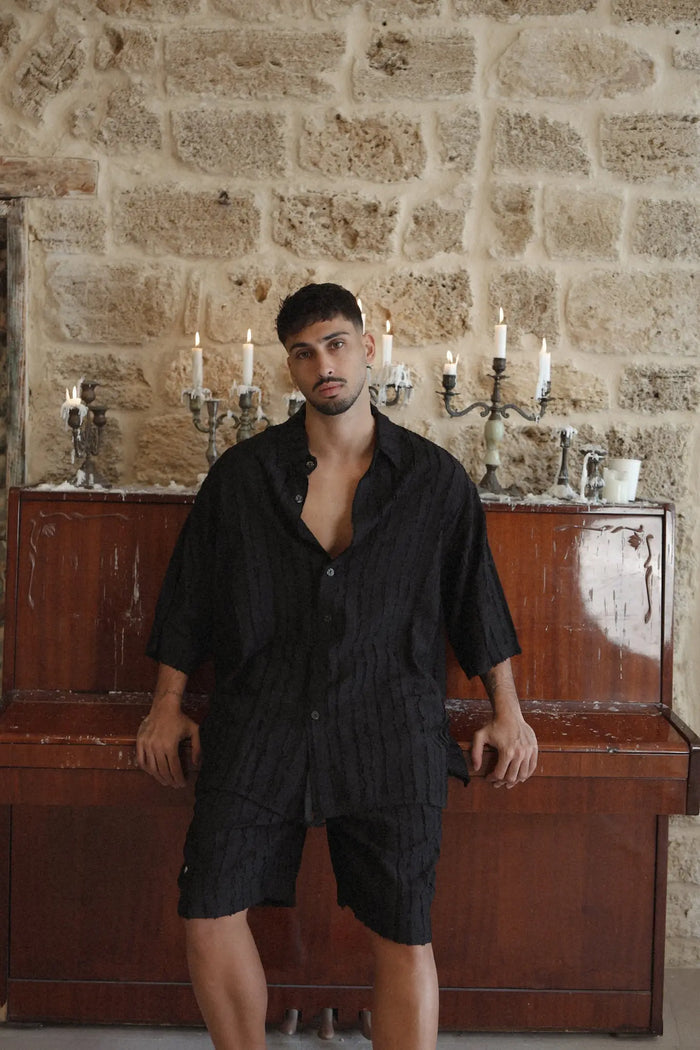 Man wearing a black RIPS Short Sleeve Button-Up Shirt and matching shorts, leaning on a piano with lit candles