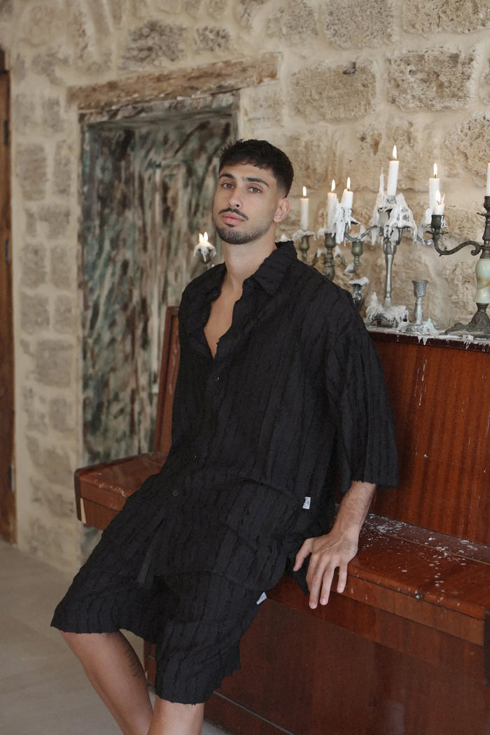 Man wearing RIPS Short Sleeve Button-Up Shirt in black, sitting on a wooden piano with lit candles in candelabras behind him