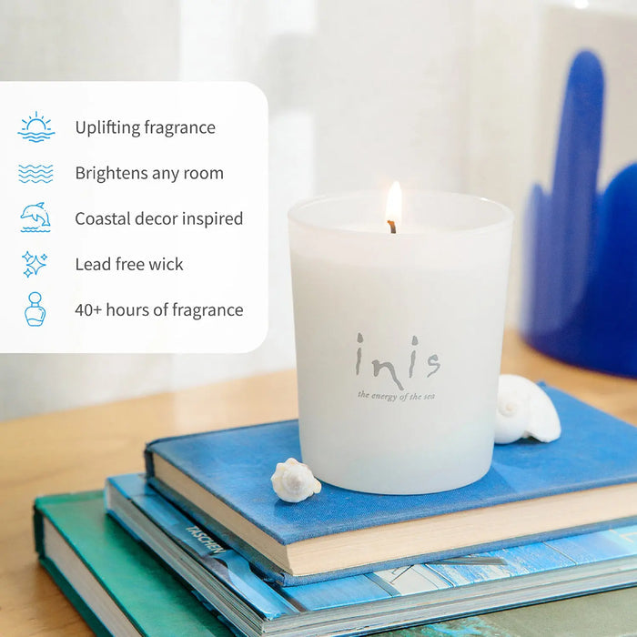 A frosted white candle labeled 'inis' sits lit on a stack of blue and green books, with small white seashells beside it and a text callout listing fragrance benefits. The scene is styled on a tabletop with soft natural light and a blurred blue decorative object in the background.