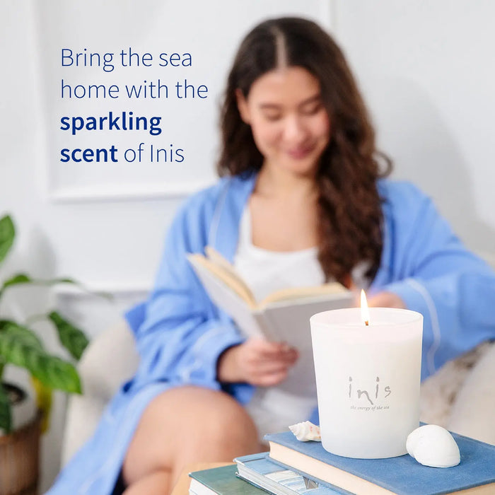 A woman in a blue robe sits on a couch reading a book, with a lit white Inis candle placed in the foreground on a table next to a small white shell. Text on the left reads, 'Bring the sea home with the sparkling scent of Inis.
