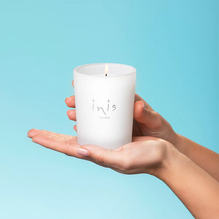 Hand holding a white candle with 'inis' branding against a light blue background