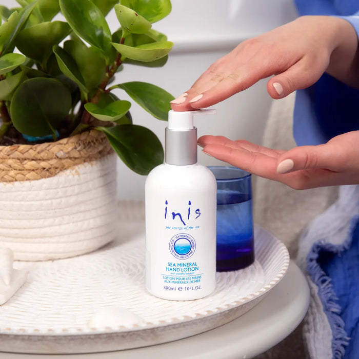 Person using a bottle of 'inis' hand lotion on a table with a plant in the background