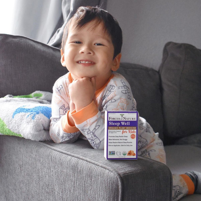 Young child in pajamas smiling on a gray couch with a Sleep Well for Kids product box on the armrest