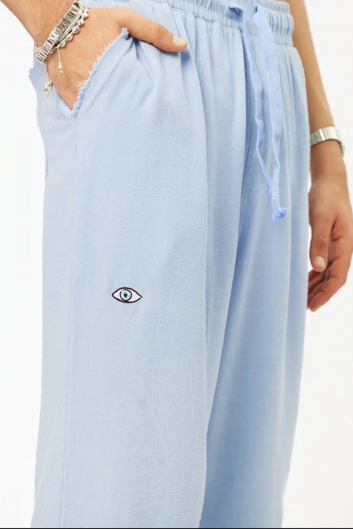 Close up of sky blue SOFT SL Linen Pants with a small embroidered eye on the left leg and a hand in the pocket