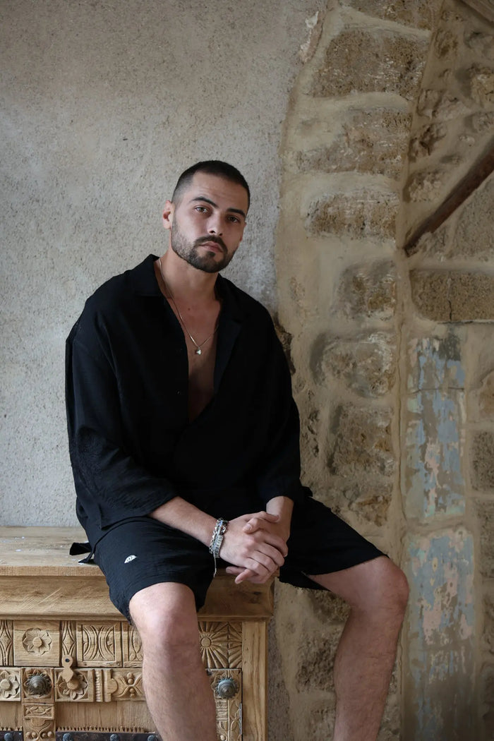 Man wearing a black SPARKS 3/4 Sleeve Button-Up Shirt, black shorts, and a silver necklace, sitting on a carved wooden chest