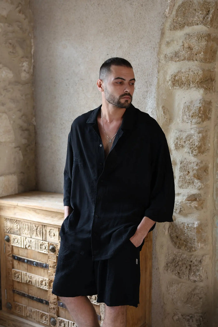 Man wearing a black SPARKS 3/4 Sleeve Button-Up Shirt and black shorts, standing next to a carved wooden cabinet