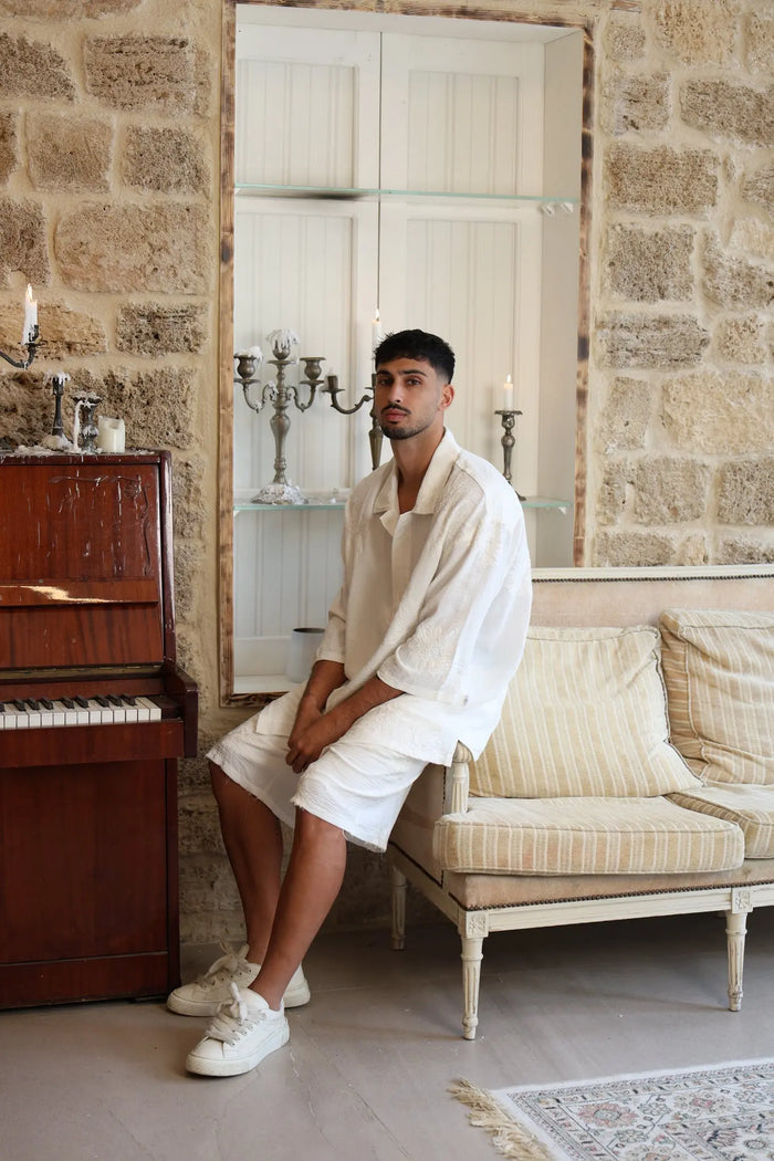 Man wearing a white SPARKS 3/4 Sleeve Button-Up Shirt and white shorts, sitting on a sofa next to a piano