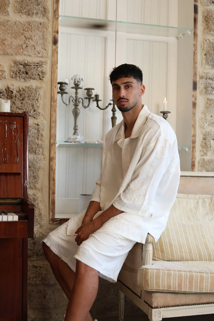 Man wearing a white SPARKS 3/4 Sleeve Button-Up Shirt and white shorts, sitting on a sofa next to a piano