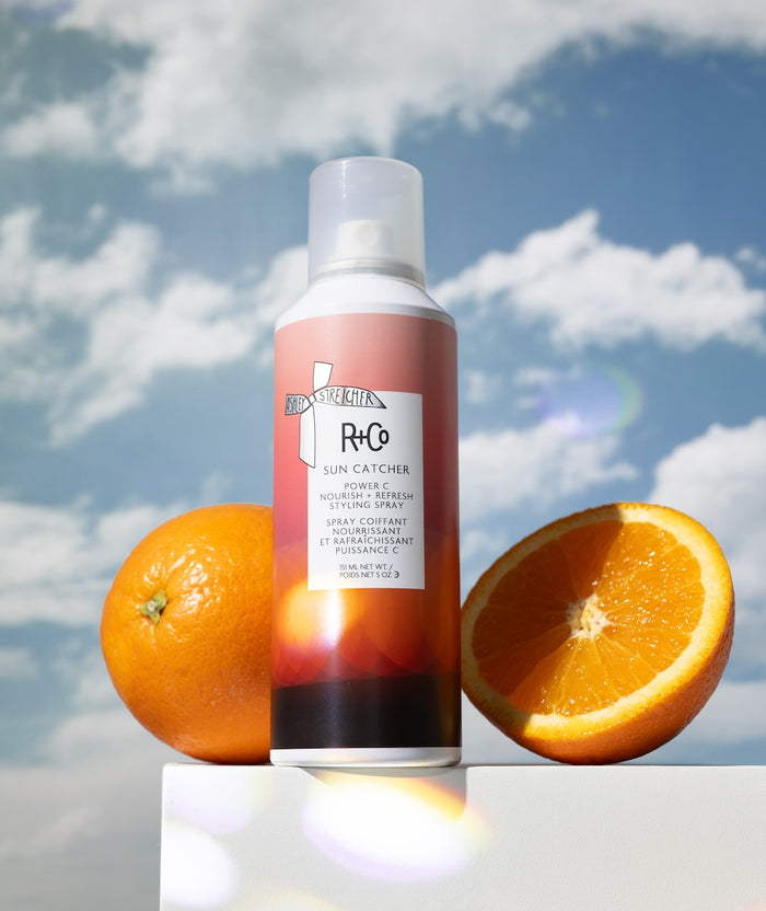 R+Co Sun Catcher hair spray bottle with oranges against a cloudy sky background
