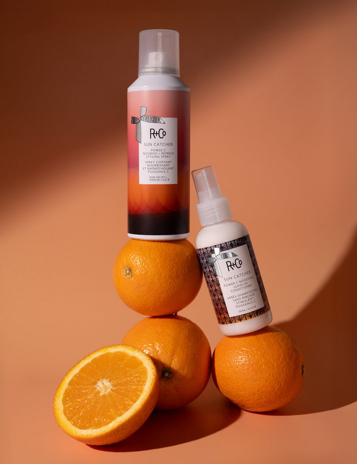 Two bottles of R+Co hair products on oranges with a warm brown background