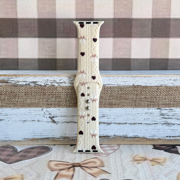 Close-up detail of Sweater Weather Sweetheart Apple Watch band showing the cream cable-knit texture, taupe bow print, and brown heart accents.