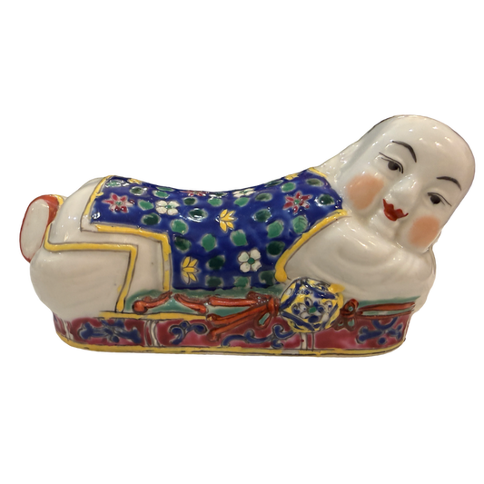 A ceramic figurine of a reclining figure in traditional East Asian-style clothing, decorated with a blue floral pattern, red and gold details, and a white face and hands, isolated on a white background.