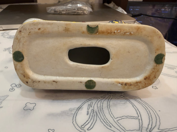 A vintage ceramic soap dish or tray with a central oval cutout, featuring a cream-colored glazed surface with green dots and brown patina around the edges, resting on a patterned tablecloth.